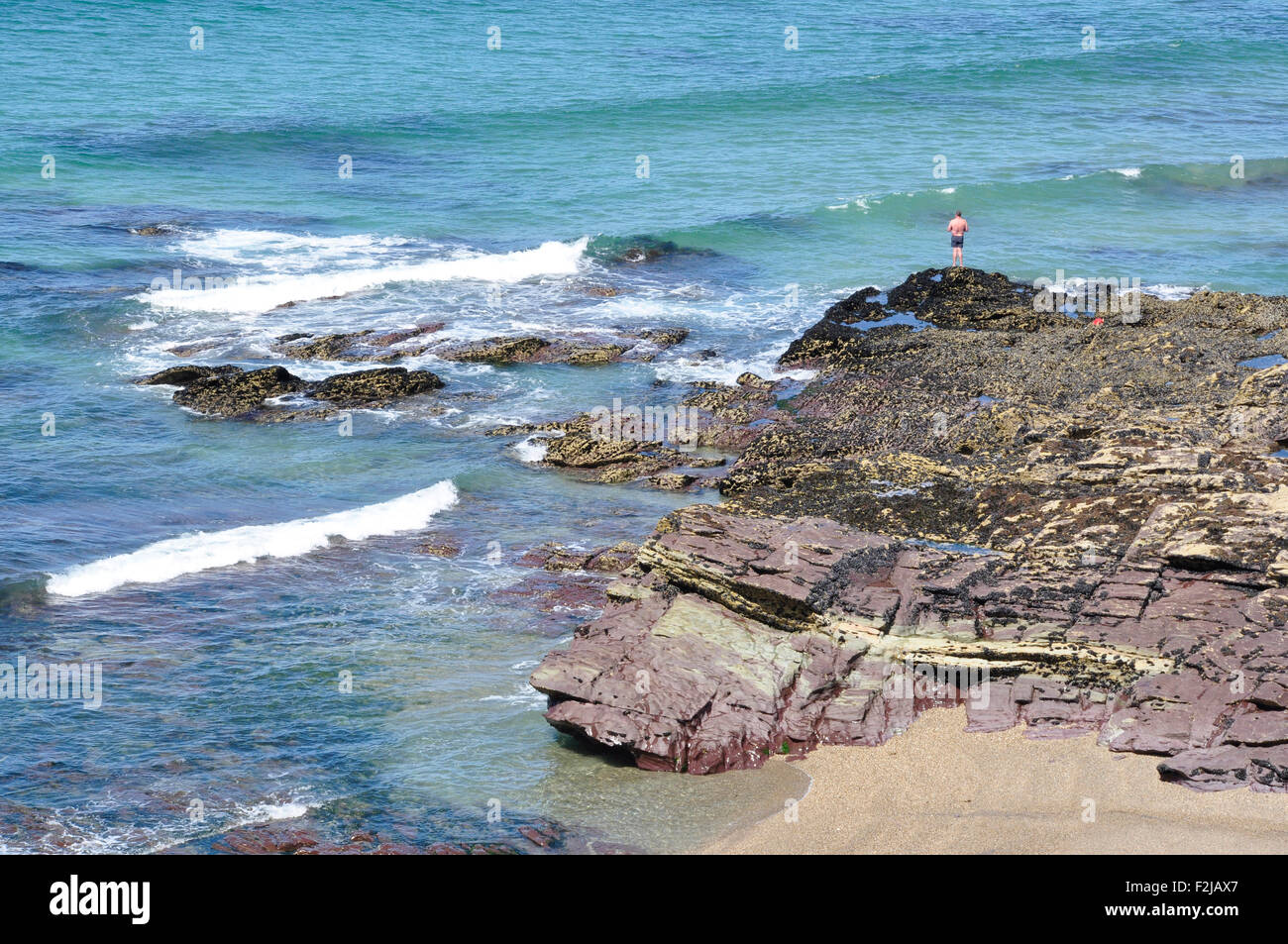 Bagnante solitaria - tiny figura appollaiato sulla roccia sopra il mare - le onde che si infrangono - blu mare - sole brillante Foto Stock