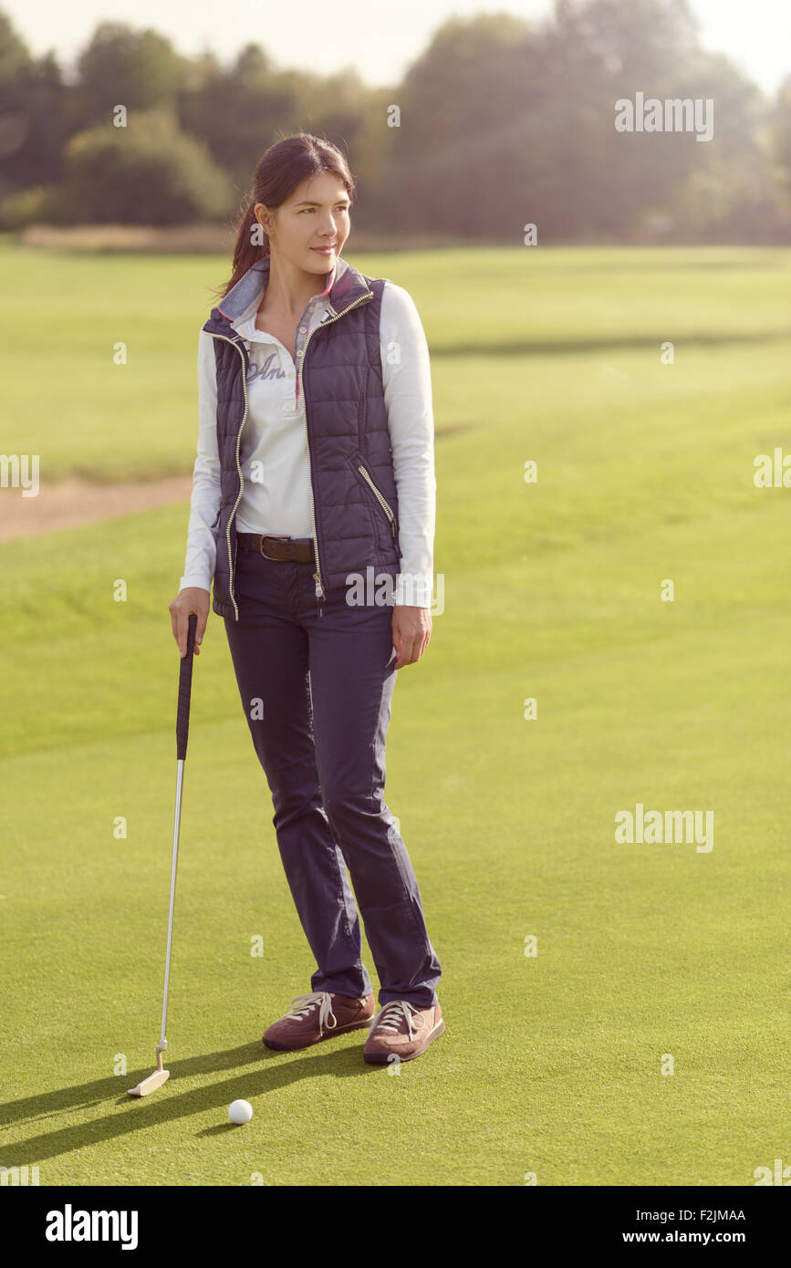 Attraente golfista femmina in piedi sul verde con il suo putter, guardando lontano con concentrazione al prossimo sciopero Foto Stock