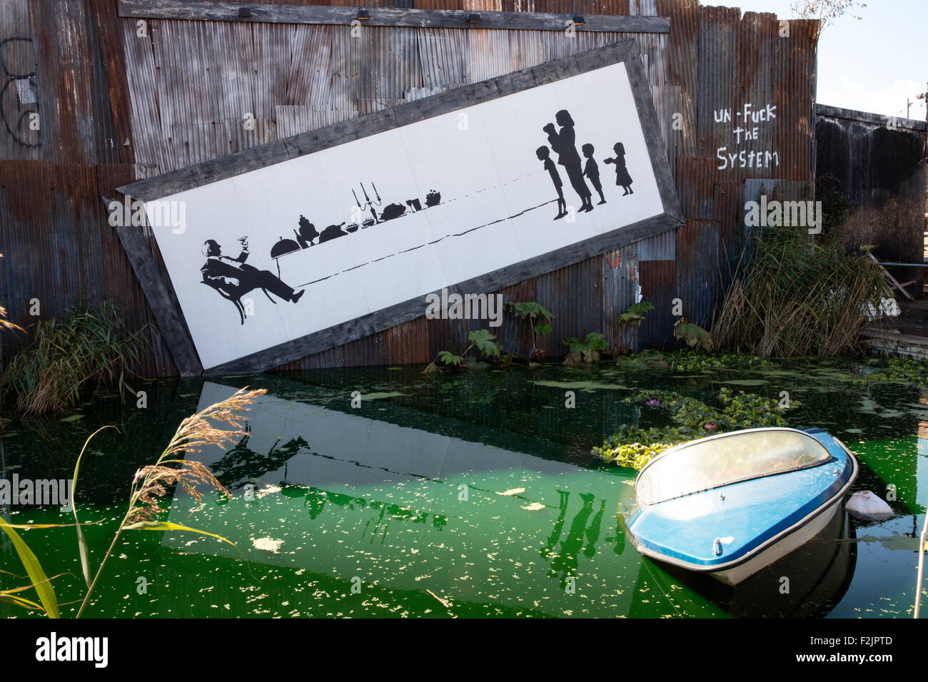 Un poster gigante di società non bilanciato da un livido verde lago in barca con barca affondata a Banksy's Dismaland Weston REGNO UNITO Foto Stock