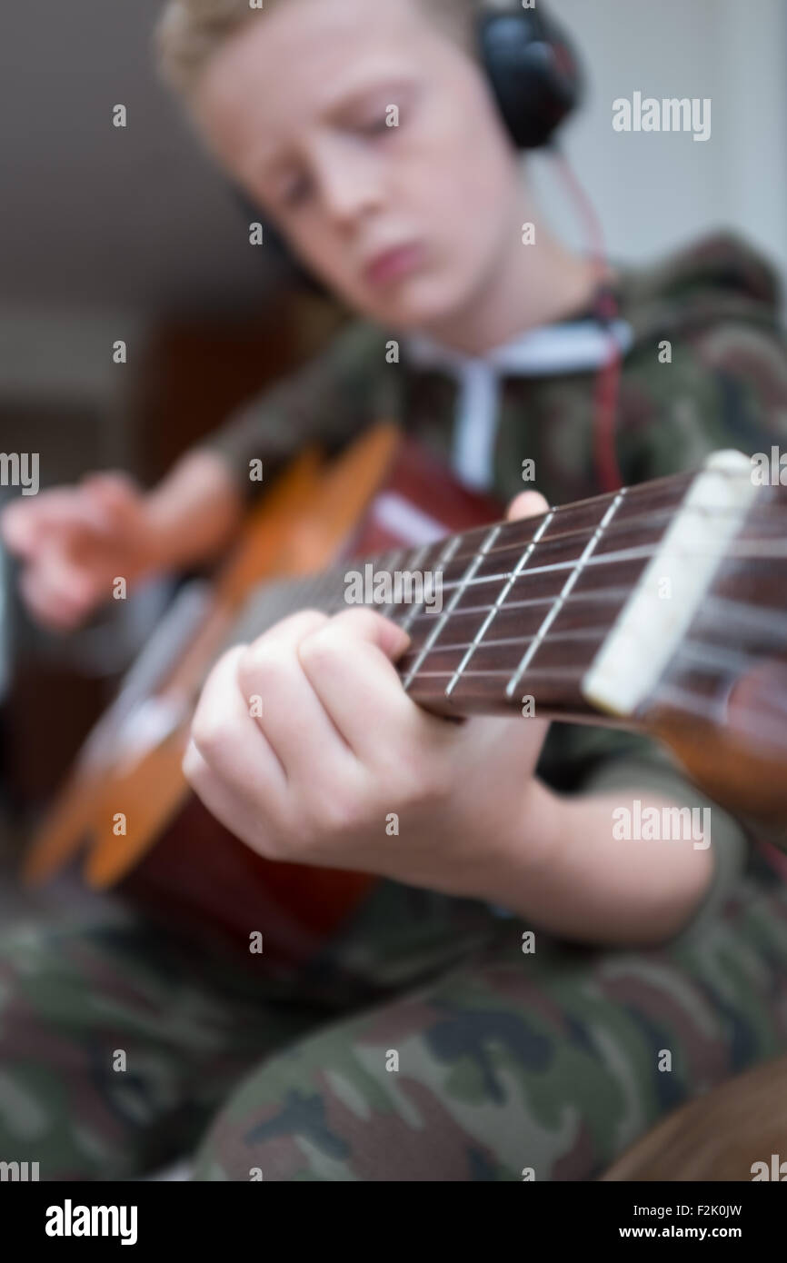 Bionda e giovane ragazzo caucasico 11 anni pratica la chitarra acustica che indossa le cuffie Beats Foto Stock