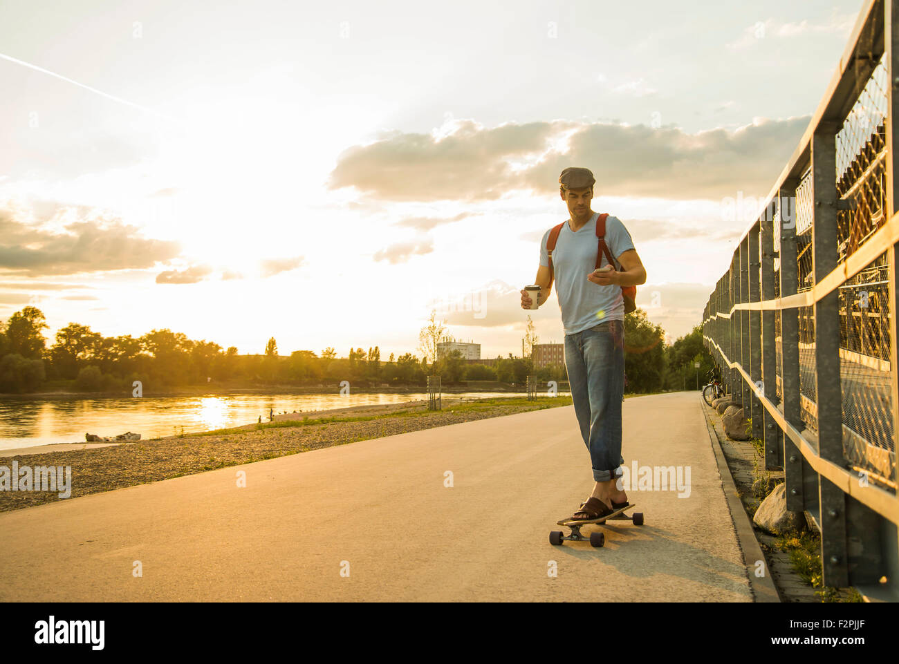 Uomo con caffè di andare e lo smartphone in piedi su skateboard nel crepuscolo della sera Foto Stock