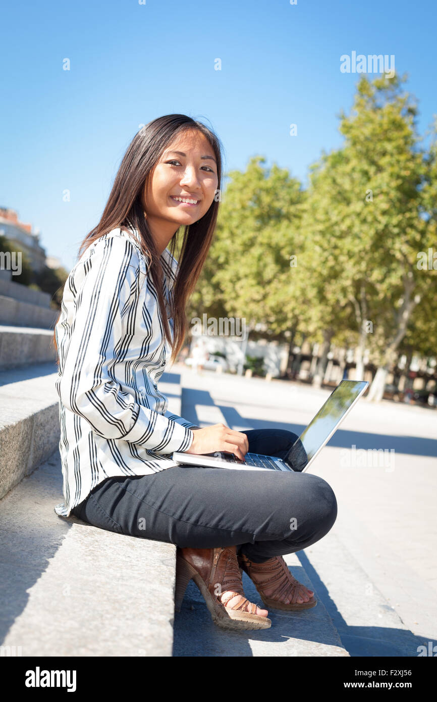 Felice ragazza asiatica usando un computer portatile seduti sulle scale in un parco Foto Stock