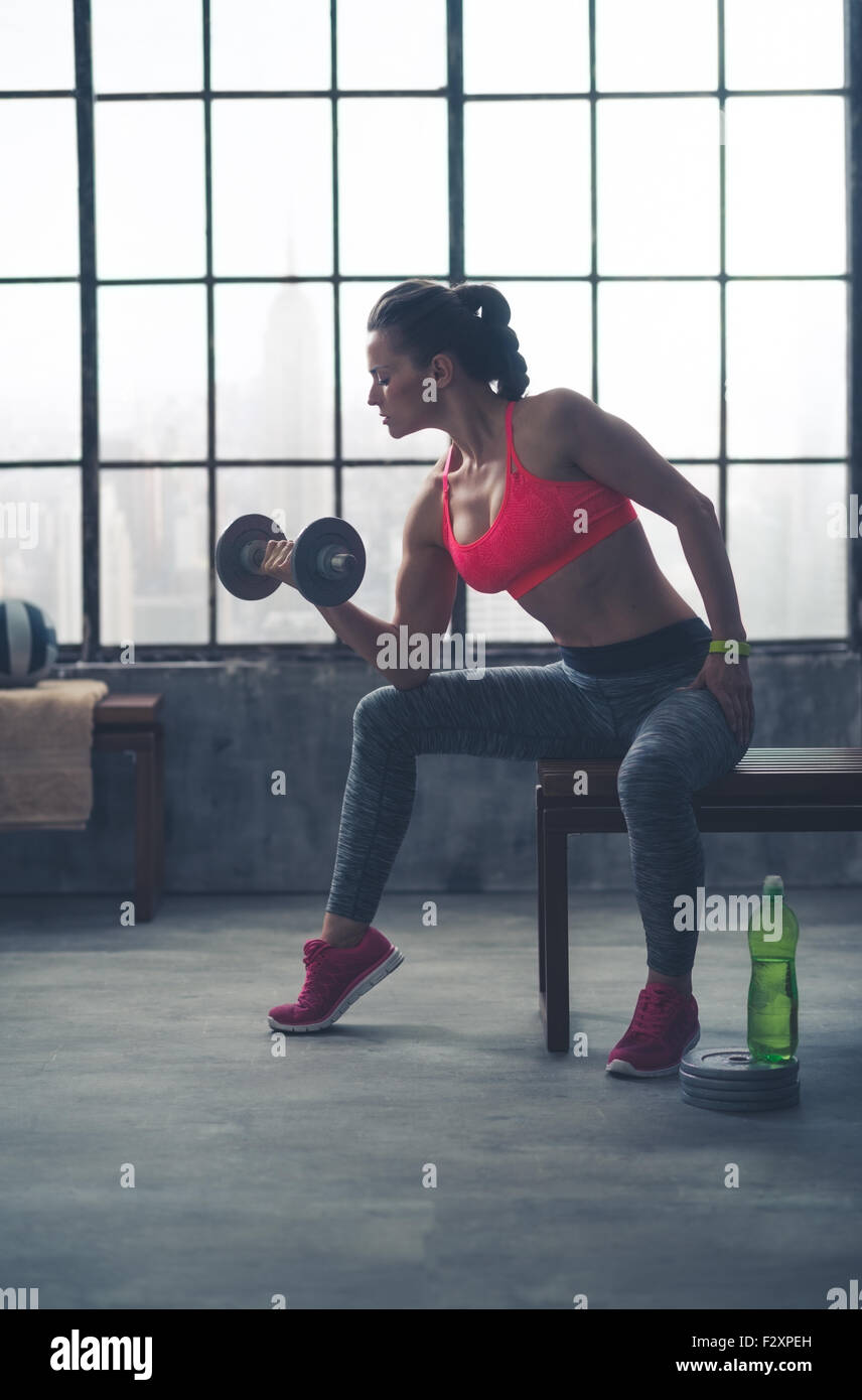 Un accoppiamento, sportivo giovane donna è seduta nel profilo su una panchina nel profilo, sollevamento pesi con una mano, mentre appoggia il suo gomito sul suo ginocchio. Foto Stock