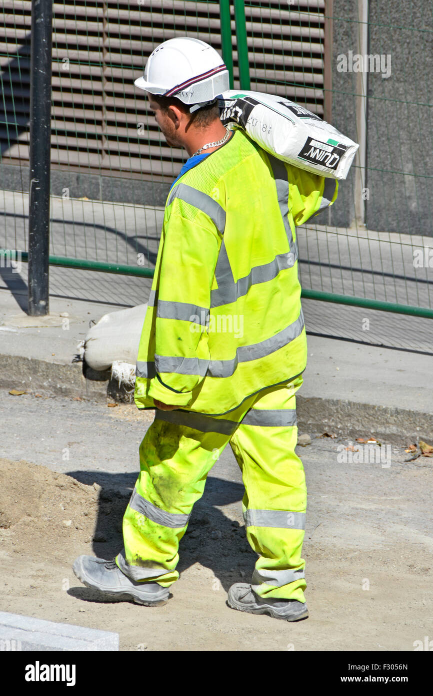 Operaio che porta un sacco di cemento sulla sua spalla indossando alta visibilità la salute & sicurezza camicia su una carreggiata sito constuction Londra Inghilterra REGNO UNITO Foto Stock
