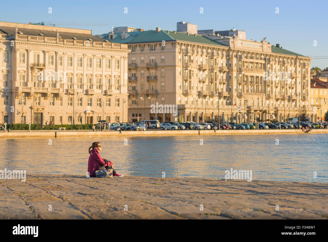 Donna che pensa da sola, guarda una giovane donna seduta da sola nel porto di Trieste e guarda il tramonto sull'Adriatico. Foto Stock