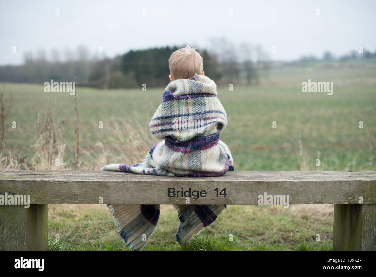 Vista posteriore di un ragazzo seduto su un banco di lavoro, avvolto in calde coperte Foto Stock