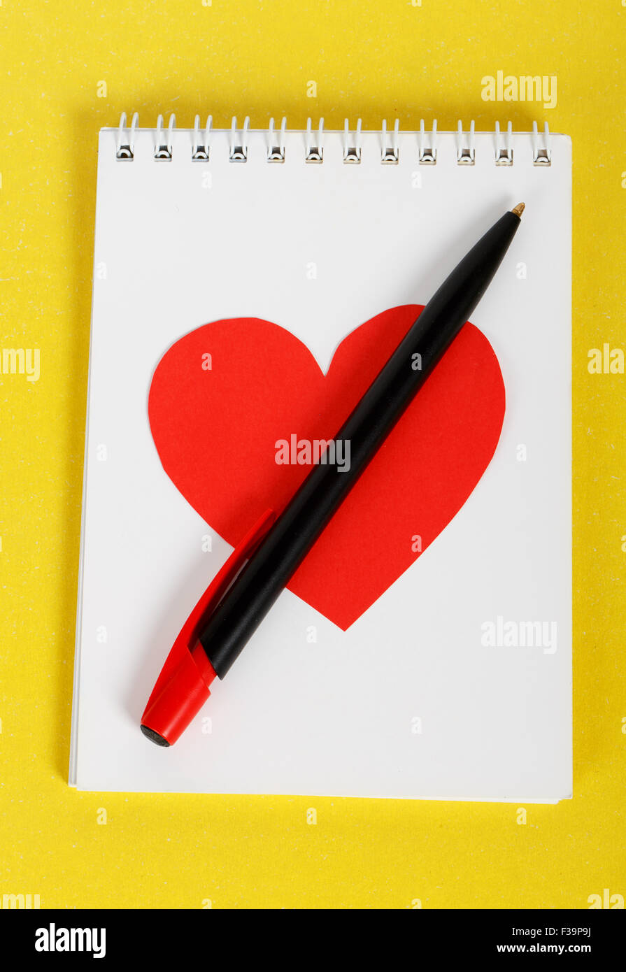 Cuore rosso con una penna nera sul notebook bianco su sfondo giallo Foto Stock