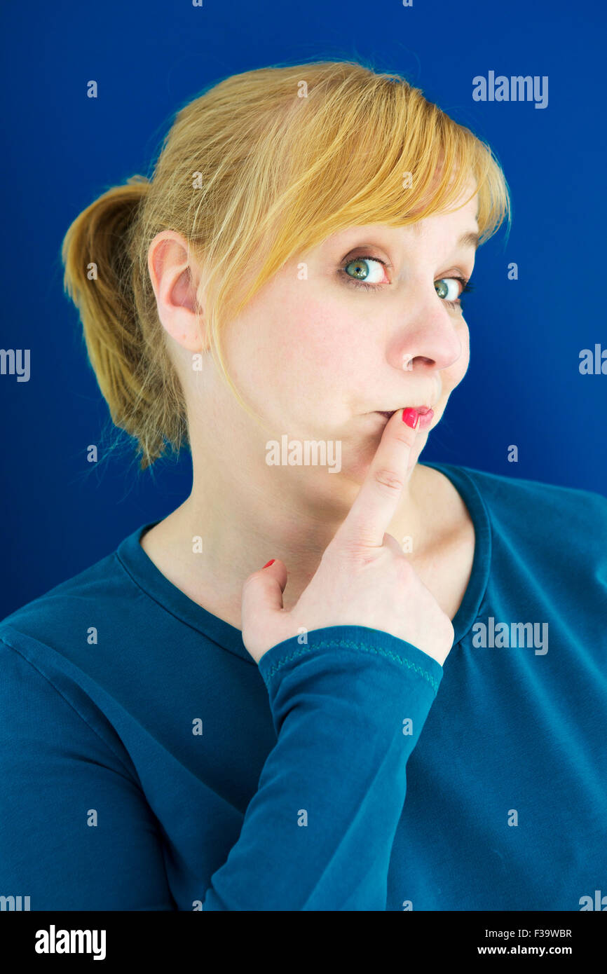 Ritratto di giovane donna bionda con sfondo blu del dito alla bocca Foto Stock