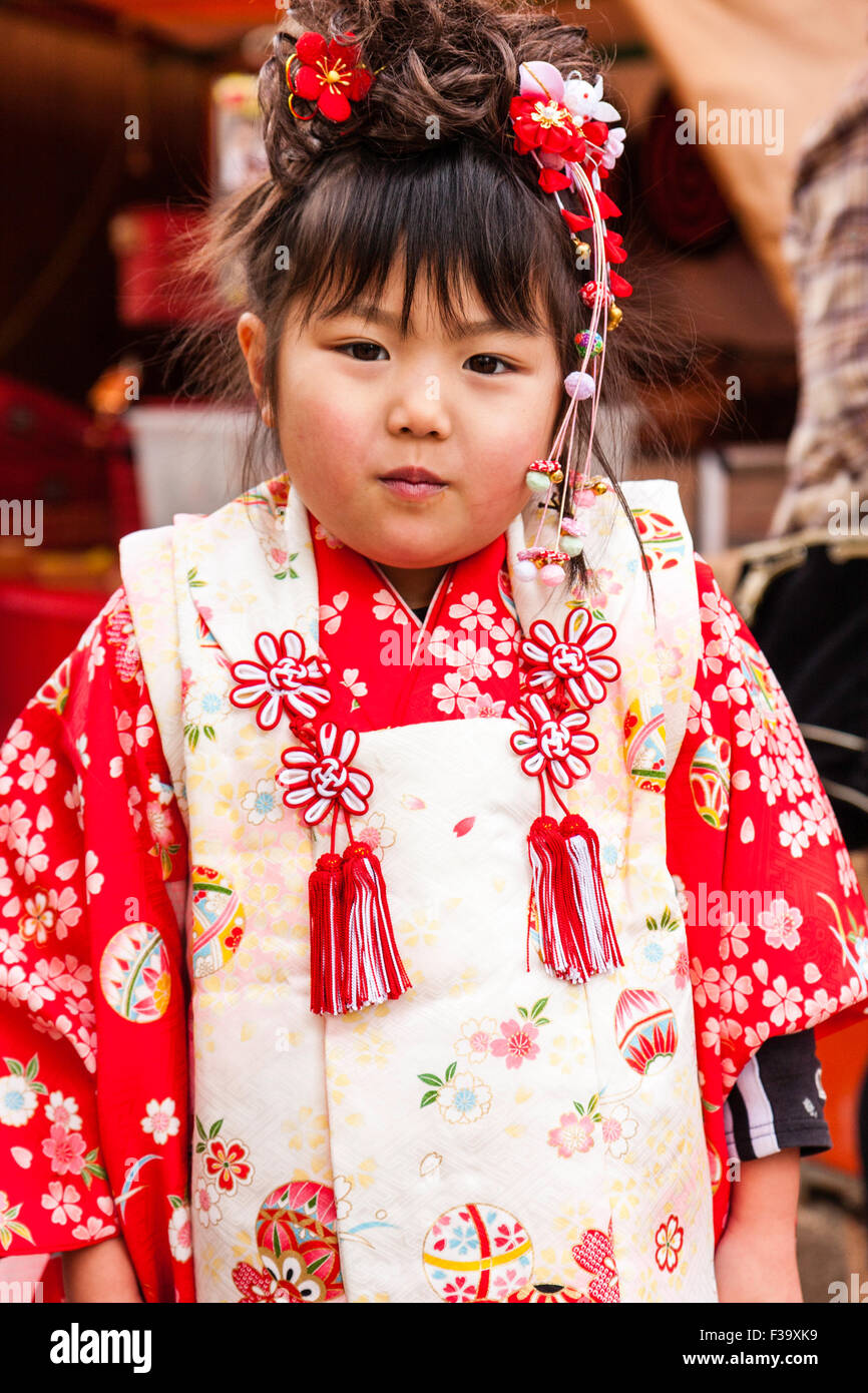 Cinque anni ragazza giapponese, leggermente al di sopra del peso con chubby guance, stando in piedi in posa per viewer indossando rosso e rosa kimono. Cheeky espressione. Foto Stock