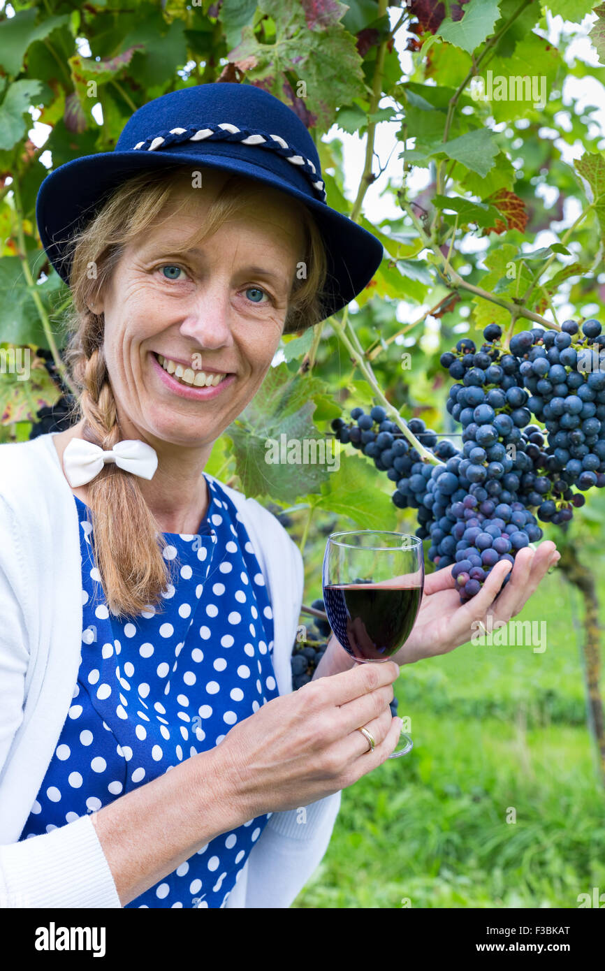 Caucasian donna di mezza età tenendo un bicchiere di vino rosso vicino a grappolo di uva blu in vigna Foto Stock