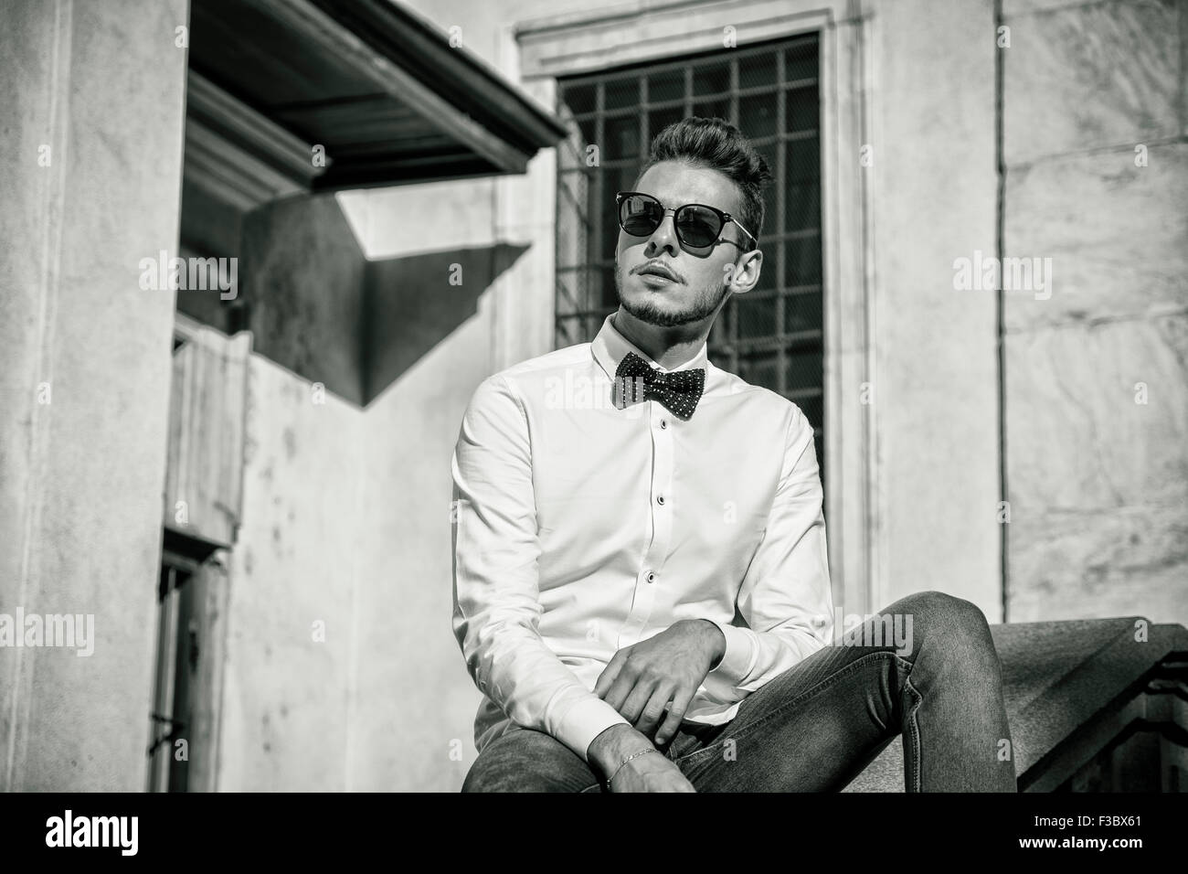 Attraente giovane uomo seduto accanto alle pareti in città, guardando lontano, indossando occhiali da sole. Mezzo corpo shot Foto Stock