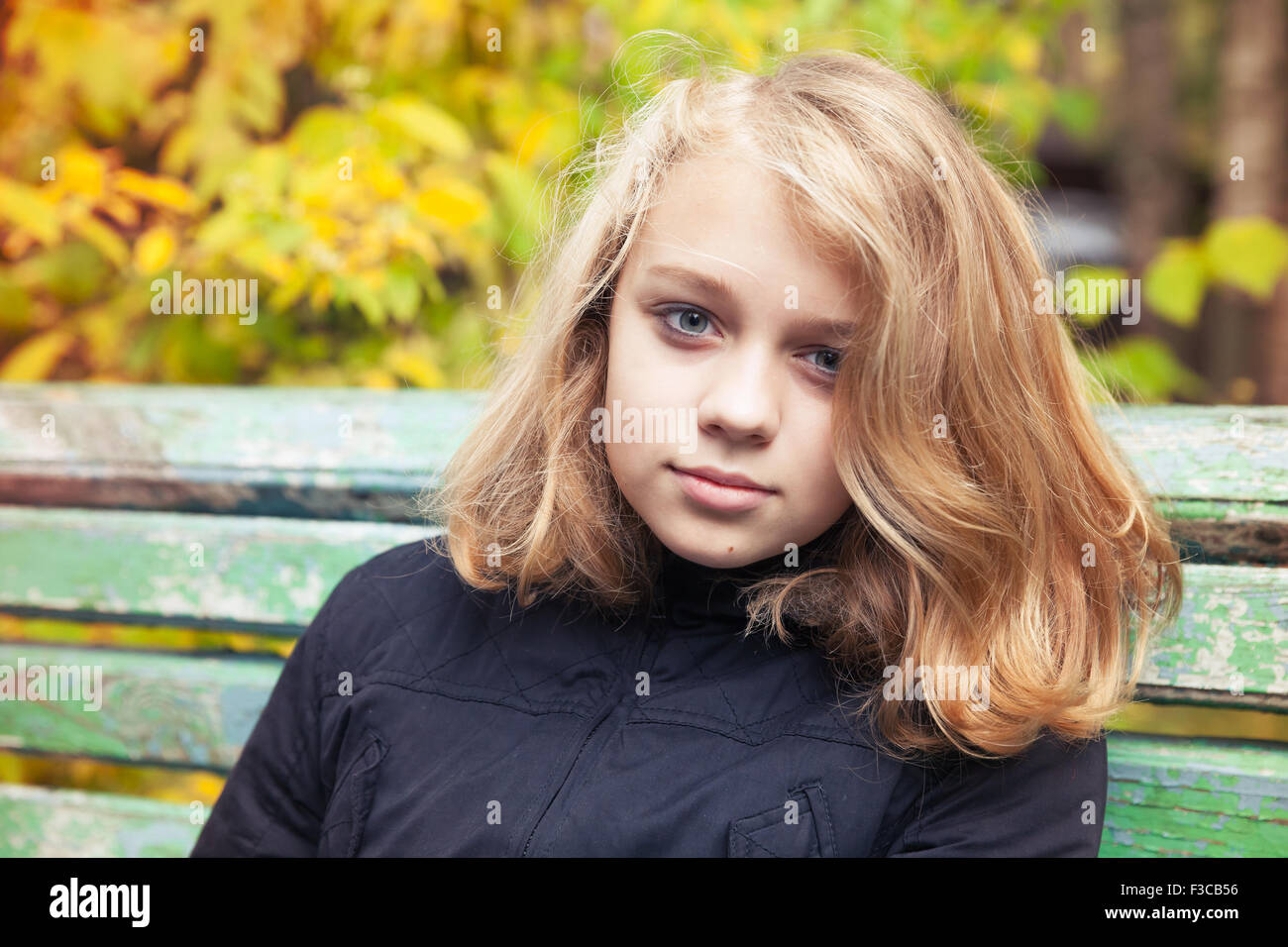 Bella bionda caucasica ragazza adolescente in giacca nera seduta sul vecchio banco verde nel parco di autunno Foto Stock