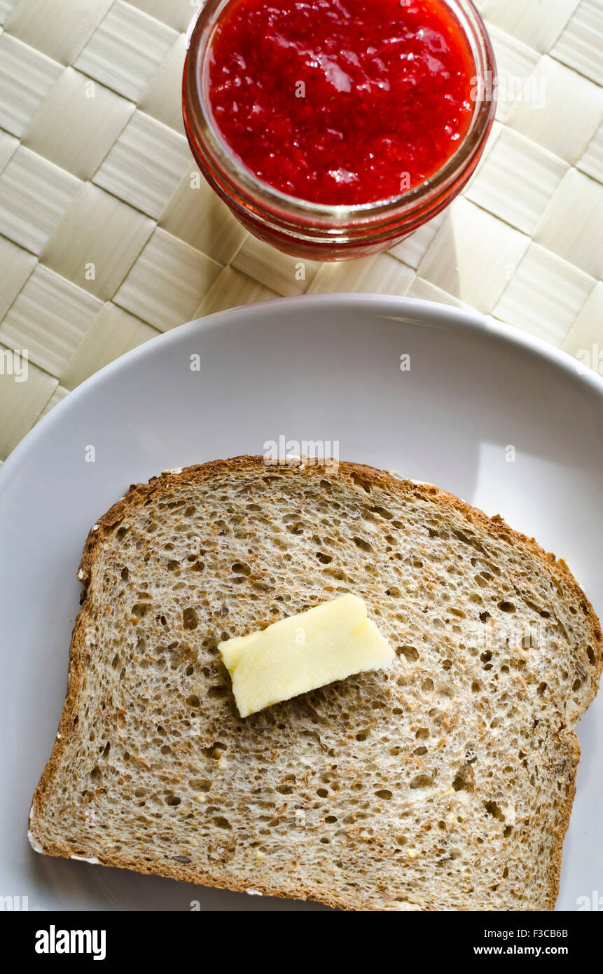 Affettare il pane integrale con un panino di burro su un piatto e un piccolo barattolo di marmellata di fragole fatta in casa. Fetta di pane tostato con burro e vasetto di conserve. Foto Stock