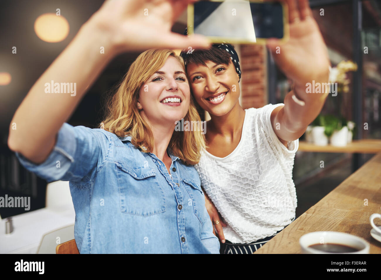 Sorridente ragazza attraente prendendo un selfie insieme come si siedono in una caffetteria gustando una tazza di caffè in posa per loro mob Foto Stock