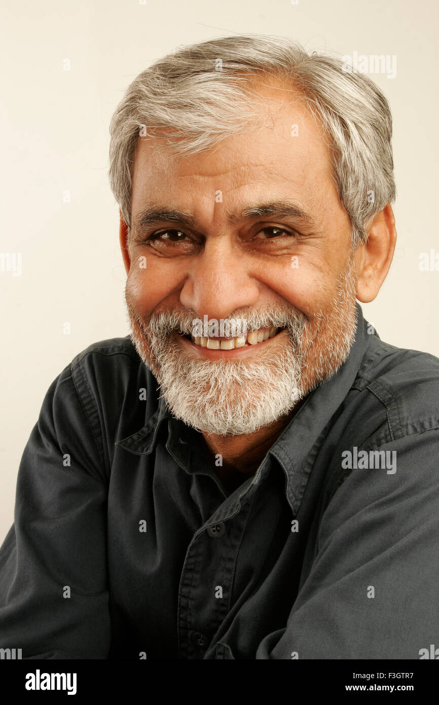 South Asian Indian vecchio uomo tardi anni cinquanta con i capelli grigi e la barba scura che indossa maglietta blu dando modesto e timido sorriso signor#340 Foto Stock