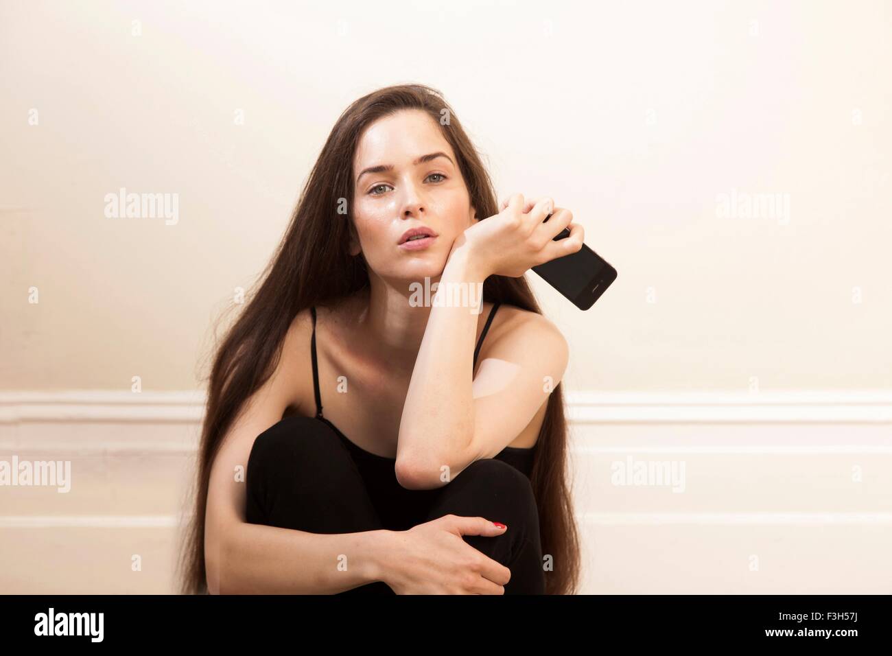 Giovane donna azienda seduta dello smartphone abbracciando le ginocchia guardando la fotocamera Foto Stock