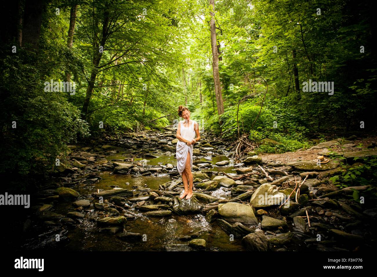 Metà donna adulta a piedi nudi sull'alveo fluviale roccioso spremitura di acqua dalla coulisse di abito bianco Foto Stock