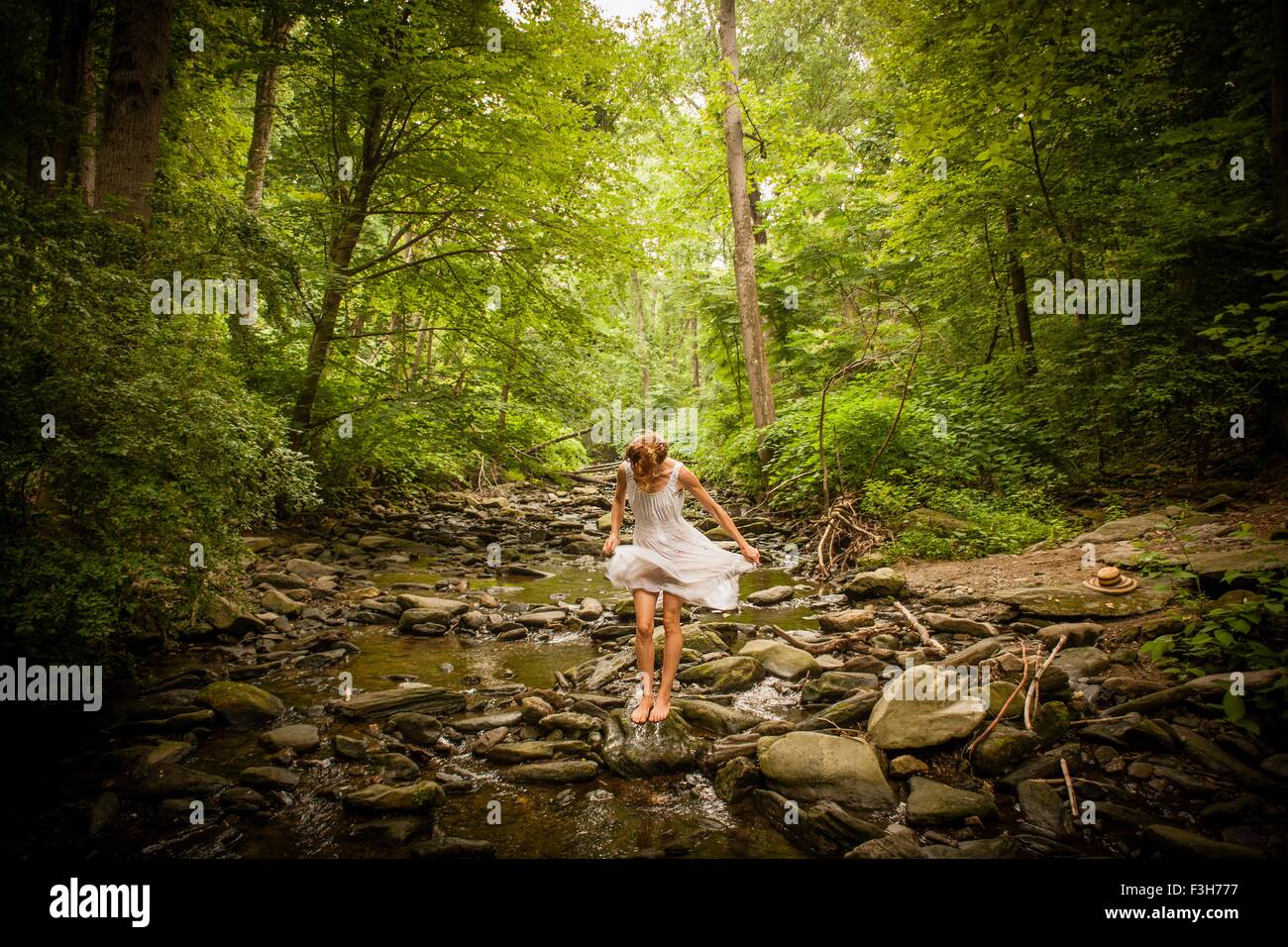 Metà donna adulta a piedi nudi sull'alveo fluviale roccioso fa roteare coulisse di abito bianco, guardando verso il basso Foto Stock