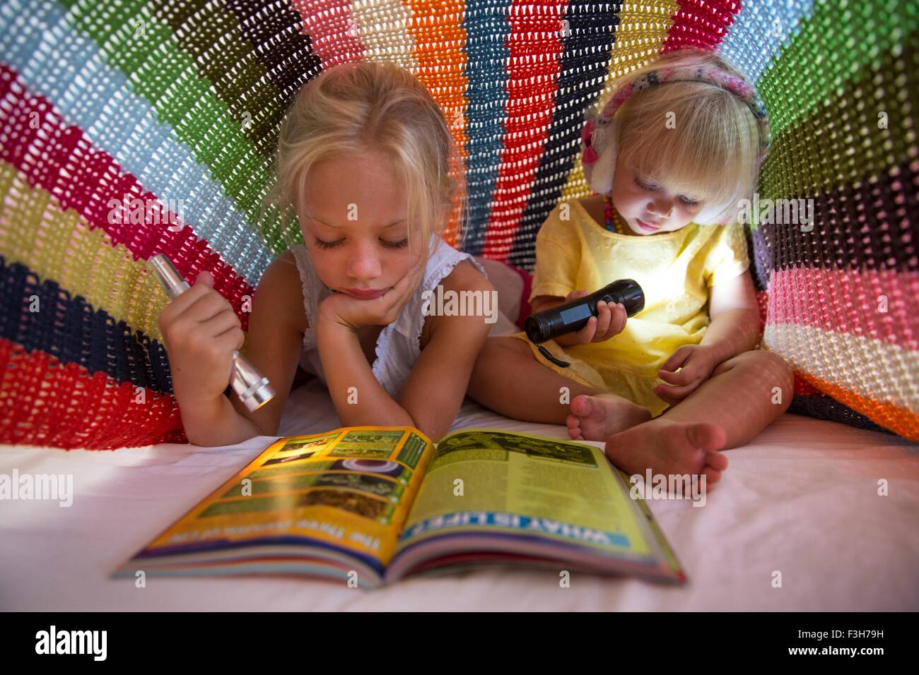 Ragazza e la sorella toddler giacente sul letto che si nasconde sotto coperta e utilizzando la torcia per leggere magazine Foto Stock