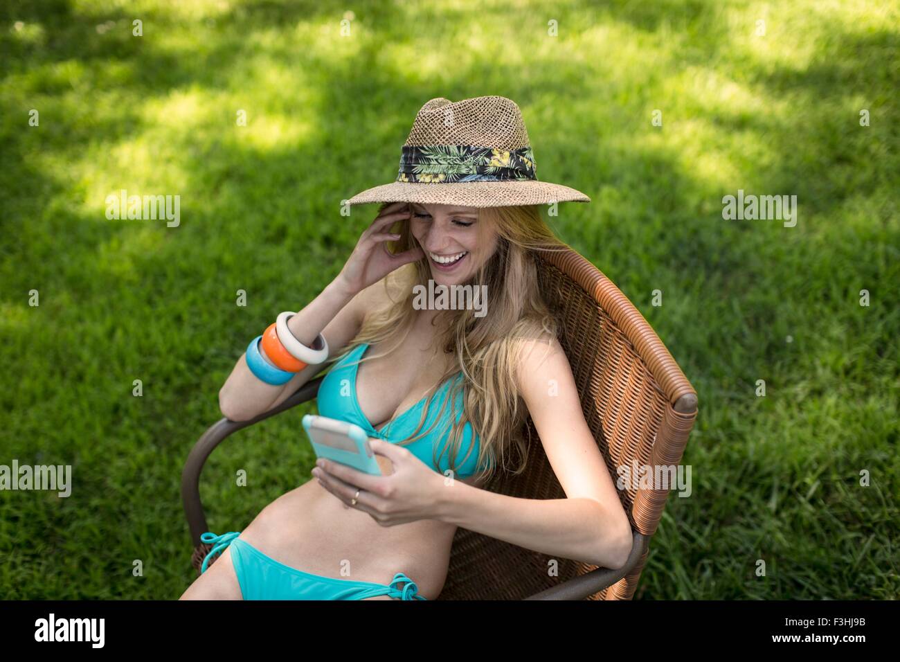 Giovane donna che indossa un bikini e cappello la lettura di testi di smartphone in giardino Foto Stock