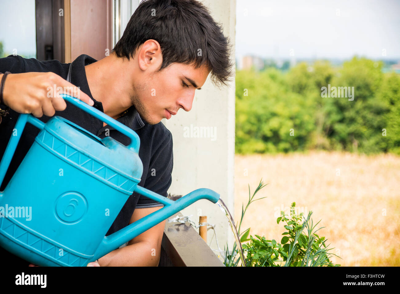 Attraente giovane uomo su Appartamento balcone Impianti di irrigazione nella casella dal blu Annaffiatoio sulla giornata di sole con il campo in background Foto Stock
