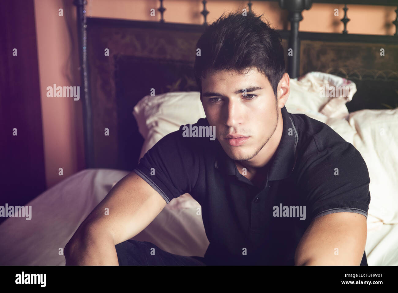 Bel giovane uomo seduto sul suo letto con espressione seria, fissando nel vuoto con uno sguardo vuoto Foto Stock
