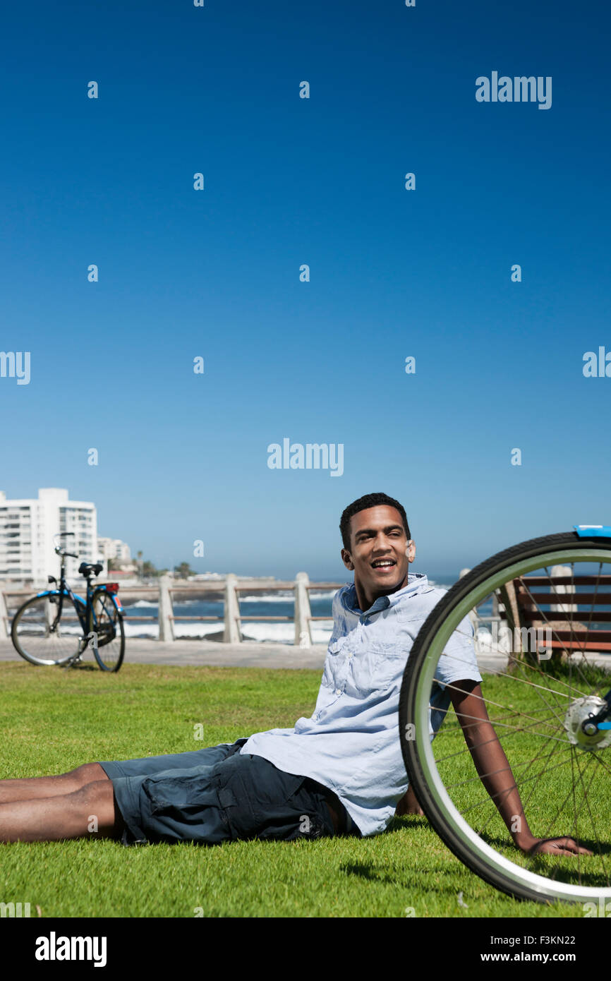 Uomo seduto sull'erba accanto alla ruota di bicicletta a Camp's Bay Beach, Città del Capo, Sud Africa Foto Stock