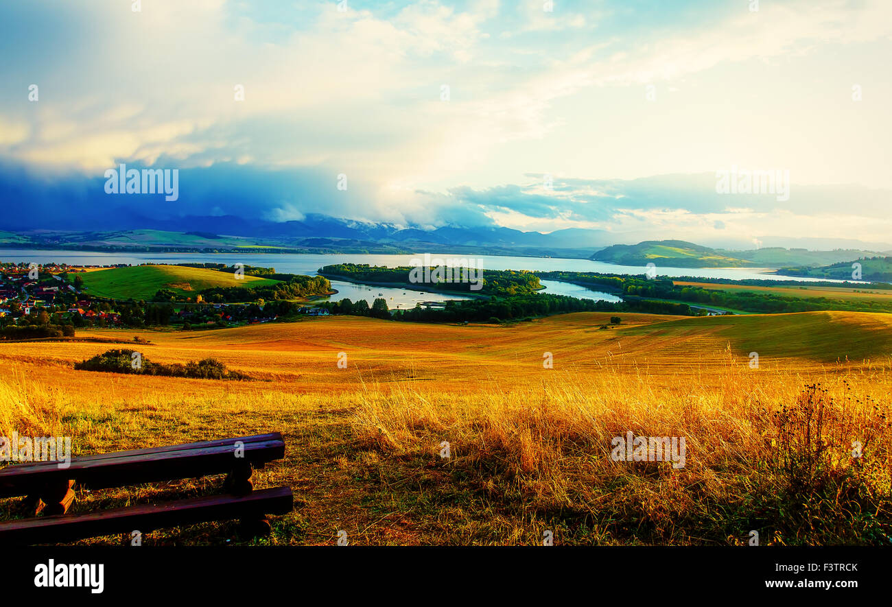 Bellissimo paesaggio. Panca in legno nel prato, affacciato sul lago e le montagne e il villaggio con un bellissimo cielo molto nuvoloso Foto Stock