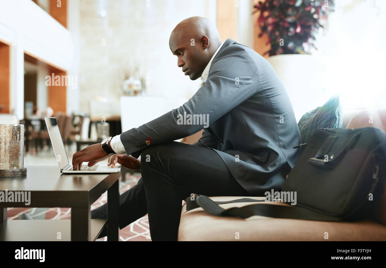Vista laterale del giovane imprenditore africani seduti sul divano a hotel lobby lavorando sul computer portatile. Foto Stock