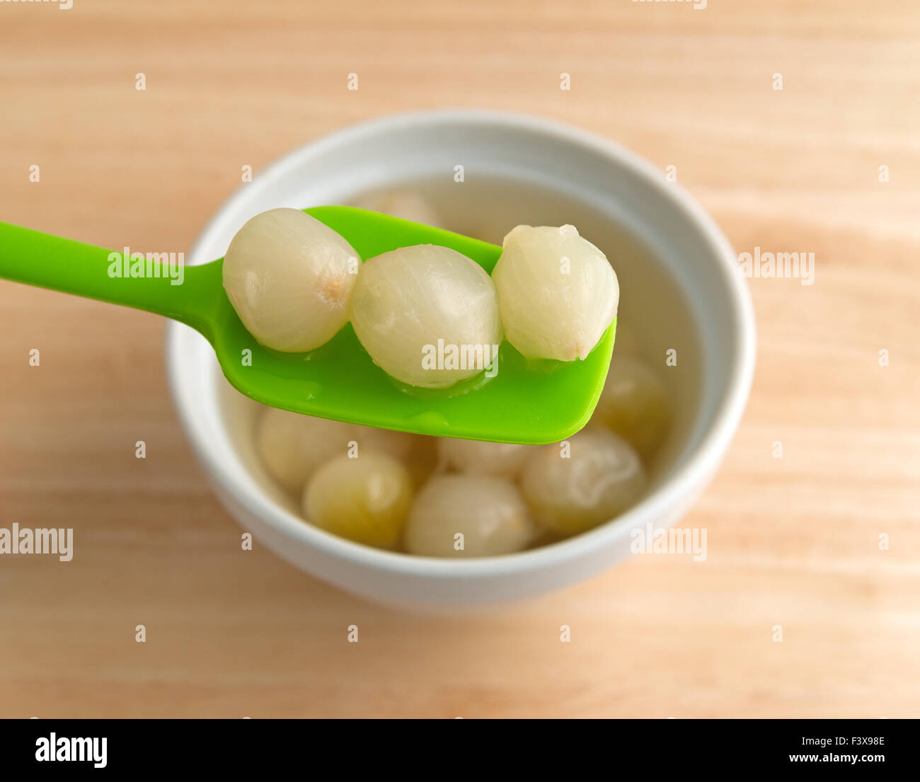 Una ciotola riempita con cotte cipolle piccole nel liquido con un cucchiaio di verde sopra con tre cipolle su una tavola di legno top Foto Stock