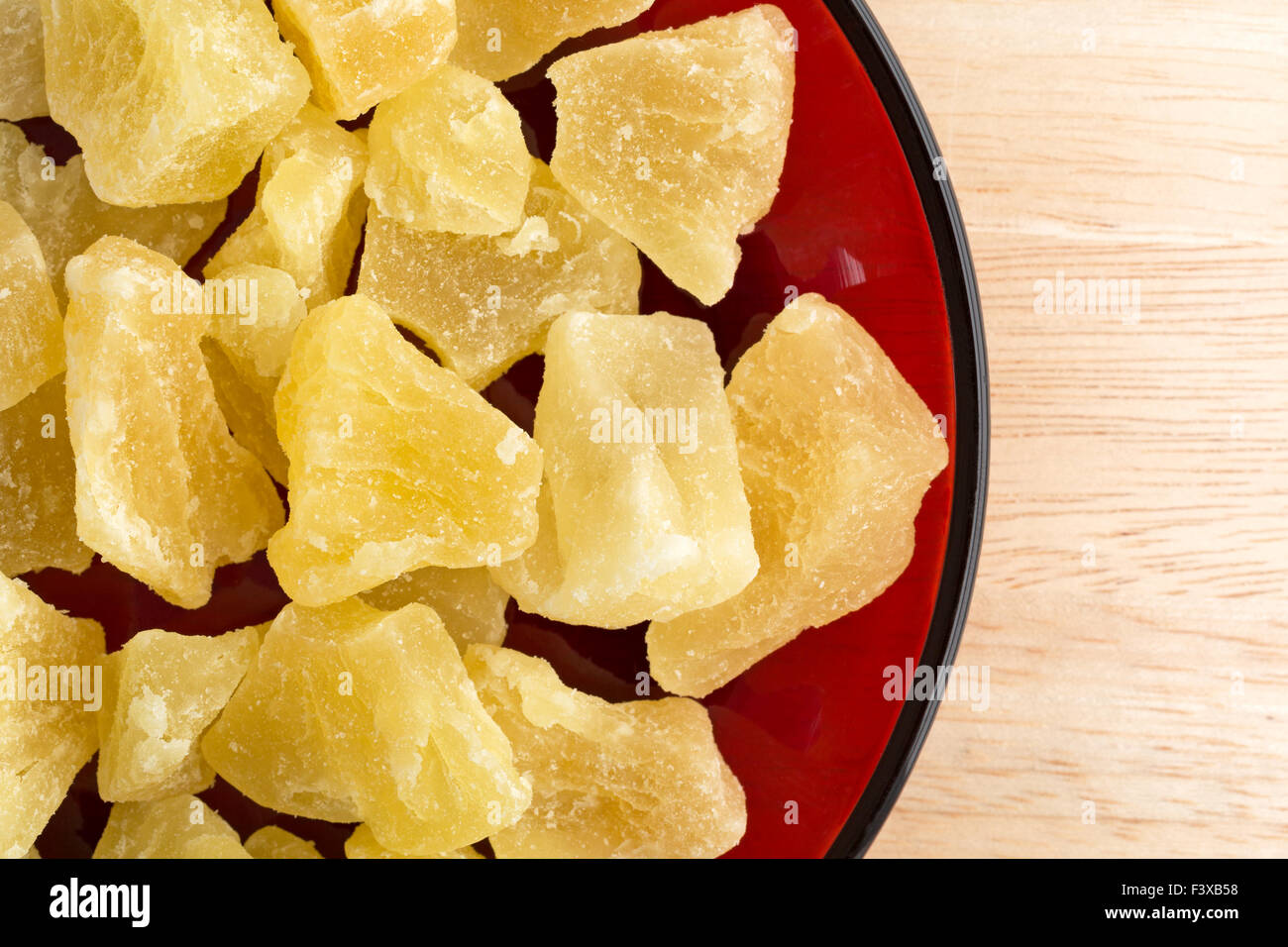 Top vista ravvicinata di un serving di essiccato confetti di ananas in pezzi su un piatto di colore rosso sulla cima di una tavola di legno alto. Foto Stock