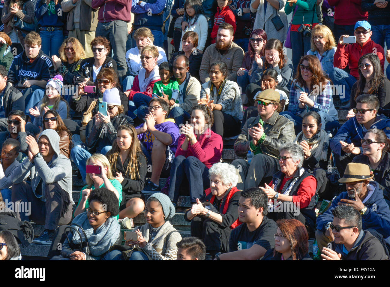Un gruppo di persone multietniche di tutte le età, ampio e diversificato, che si siedono all'esterno su bleachers in cemento che reagiscono a guardare artisti di strada che si esibiscono negli Stati Uniti Foto Stock