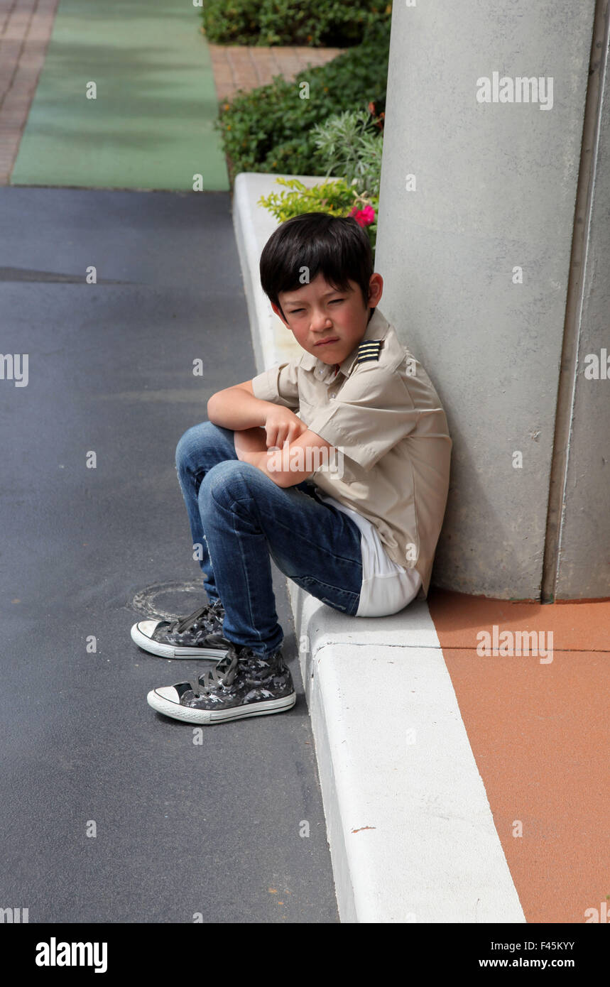 Si tratta di una foto di un burbero piccolo ragazzo che si siede sul pavimento contro una colonna o un pilastro per la strada Foto Stock