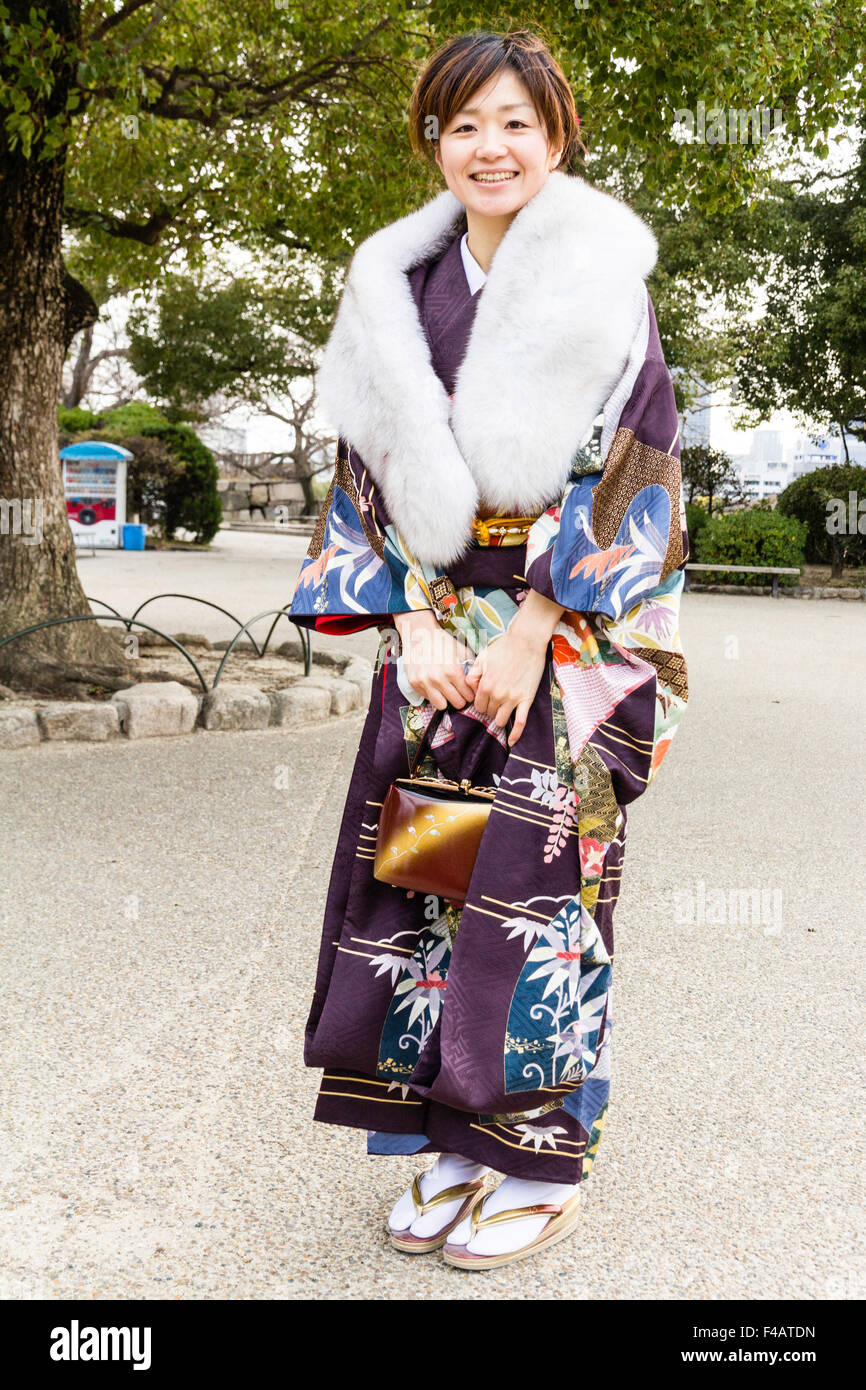 Giapponese giovane donna indossa mauve kimono e periodo invernale  Coprispalle in pelliccia. In piedi sorridente al viewer come lei in posa  per una fotografia. Detiene borsetta mediante la fascetta Foto stock -