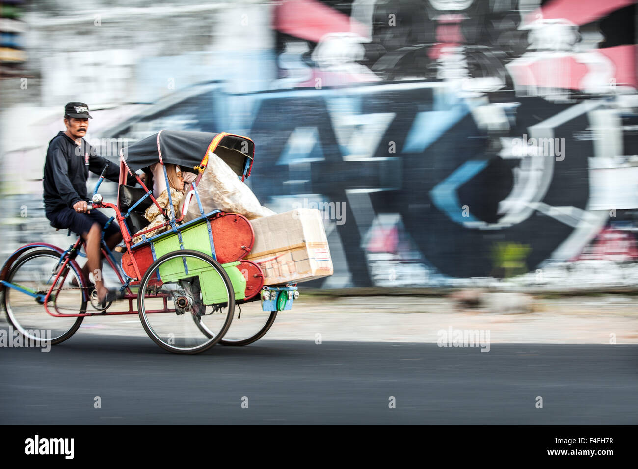 Risciò ciclo con passeggero accelerando lungo in questa fotografia panoramica Scena di strada di città indonesiane la vita con colorati graffiti sullo sfondo della parete Foto Stock