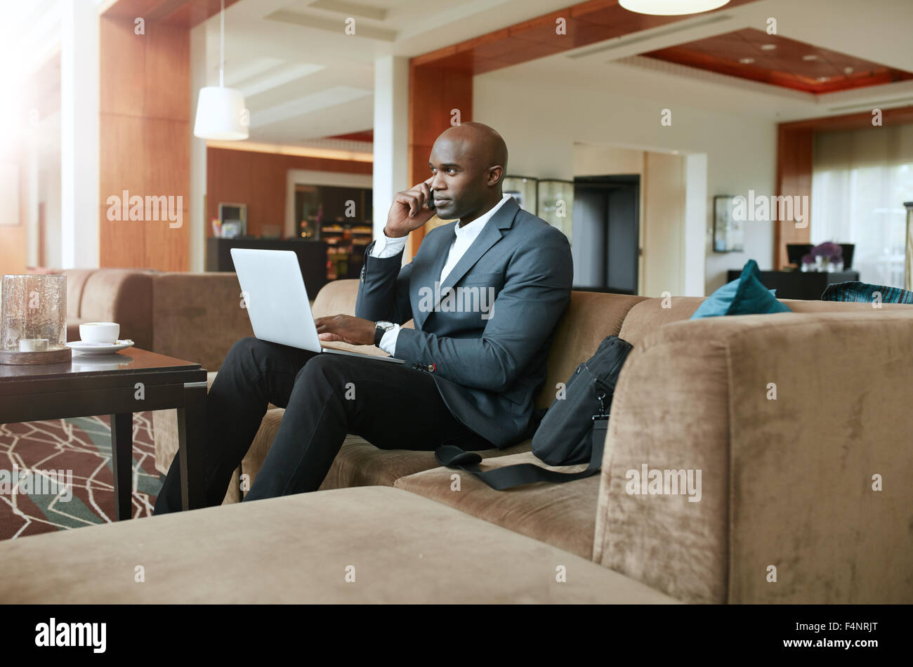 Imprenditore seduto sul divano con un laptop utilizzando il telefono cellulare. Occupato african executive maschio in attesa nella hall dell'hotel. Foto Stock