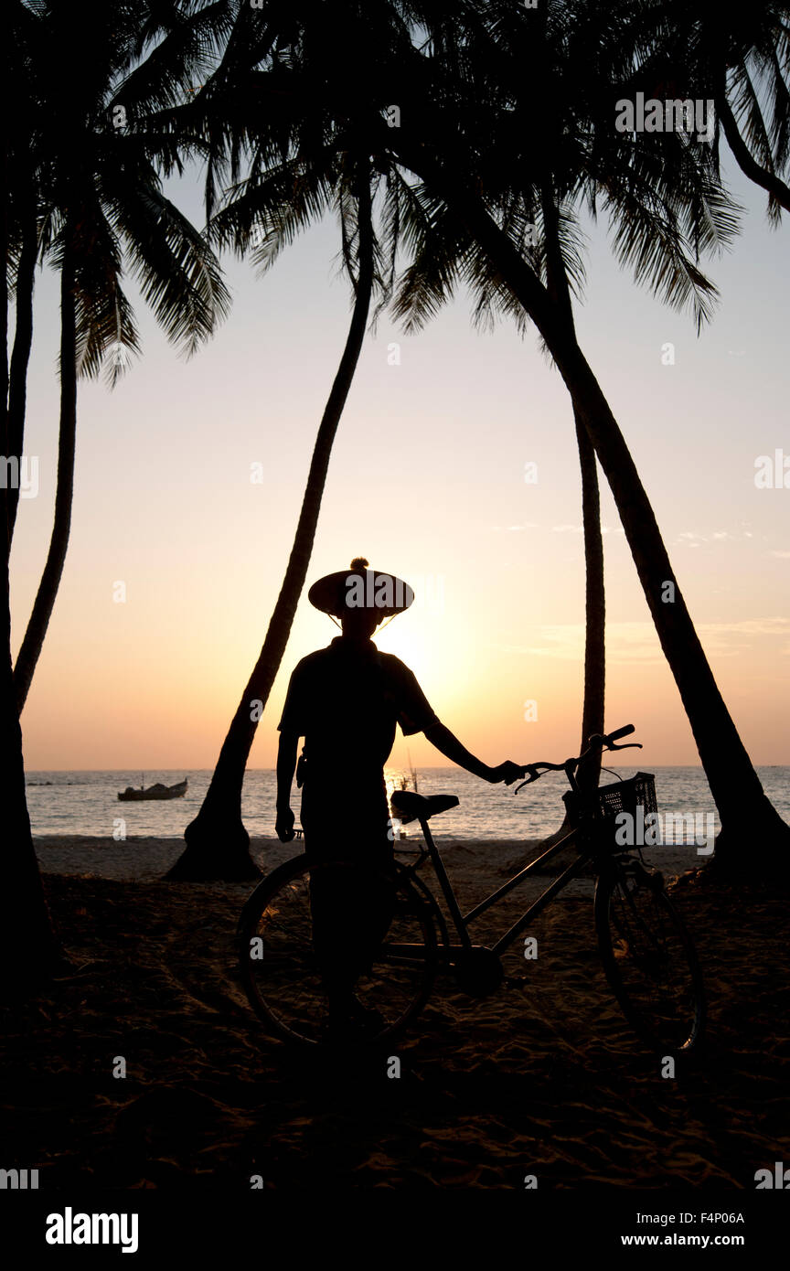 Un uomo che indossa un cappello conico con bicicletta stagliano con palme di cocco al tramonto sulla spiaggia di Ngapali Myanmar Foto Stock
