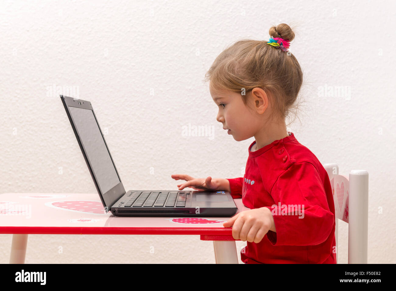 Una bionda bambina di tre anni è seduto davanti a un notebook, laptop, guardando lo schermo e utilizzando la tastiera Foto Stock