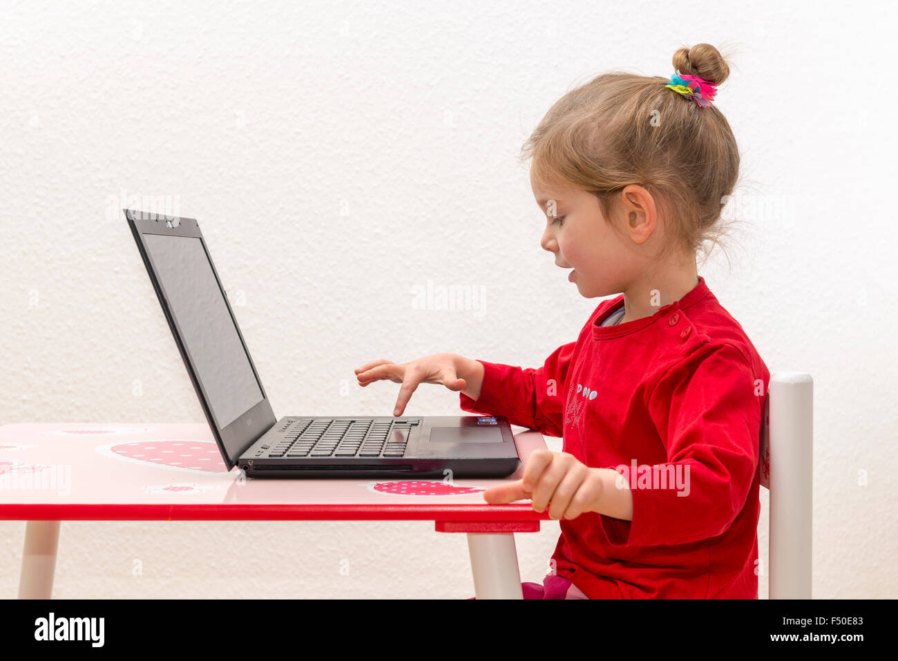 Una bionda bambina di tre anni è seduto davanti a un notebook, laptop, guardando lo schermo e utilizzando la tastiera Foto Stock