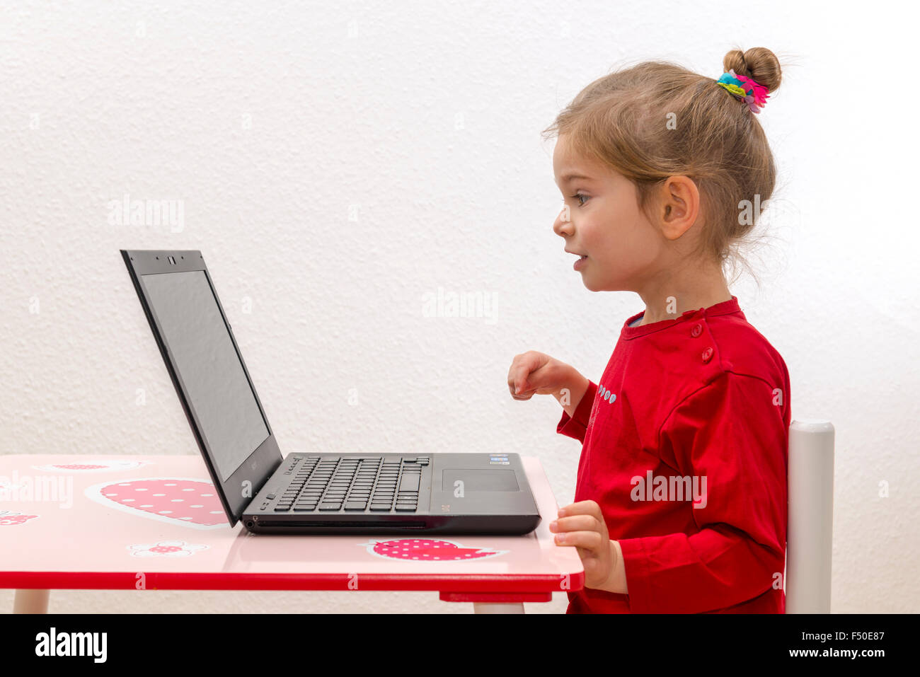 Una bionda bambina di tre anni è seduto davanti a un notebook, laptop, guardando lo schermo Foto Stock