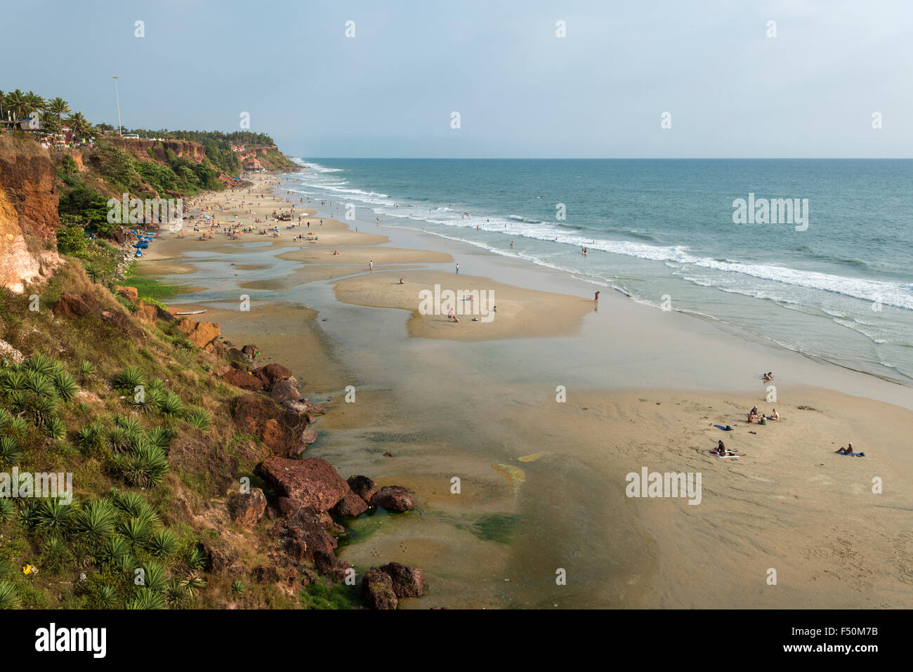 La spiaggia di Varkala con alcuni turisti bagni di sole, rosso scogliere di pietra e mare blu acqua Foto Stock