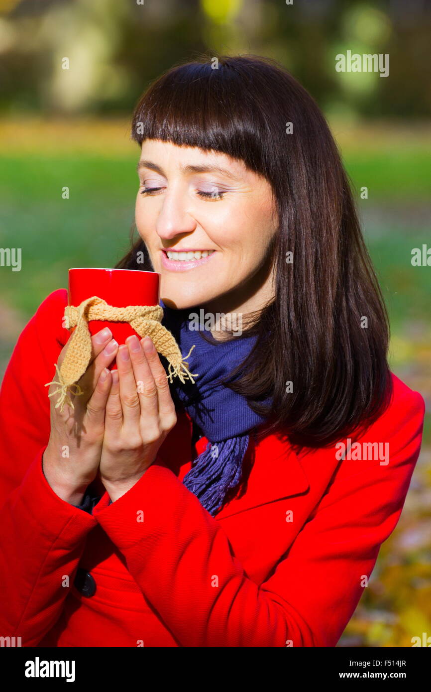 Felice e donna sorridente nel parco autunnali azienda tazza di tè caldo avvolto sciarpa di lana, autunno Foto Stock