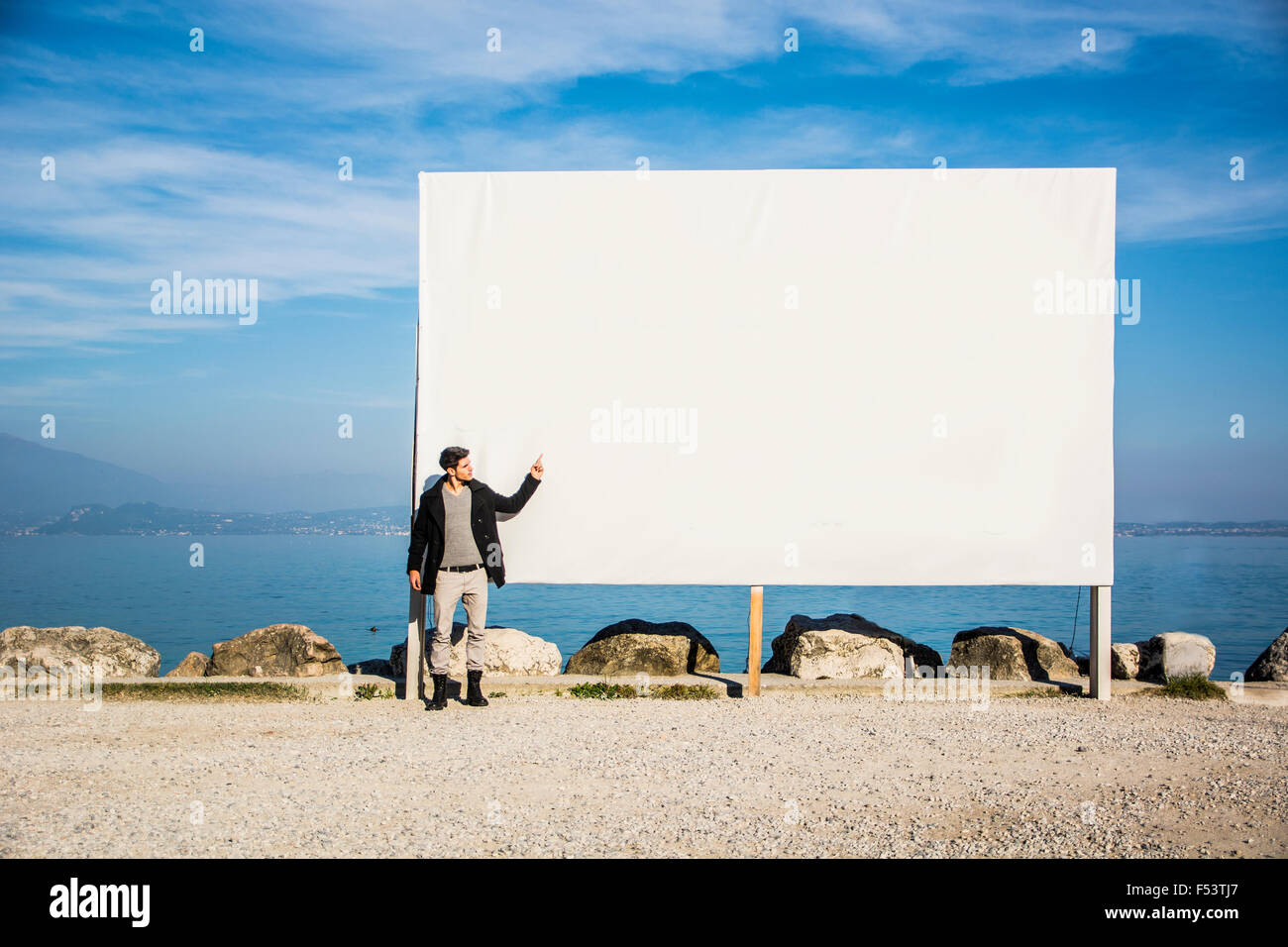 Bel giovane uomo in piedi accanto al grande vuoto tabellone bianco, display o bordo, outdoor dalla riva del lago Foto Stock