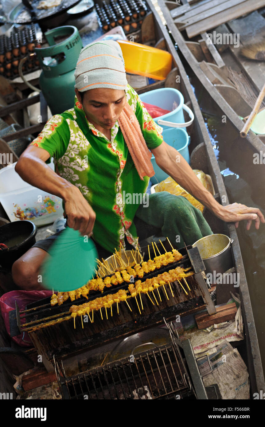 Giovane uomo grigliare gli spiedini di carne su una barca a Taling Chan mercato galleggiante di Bangkok, Tailandia Foto Stock