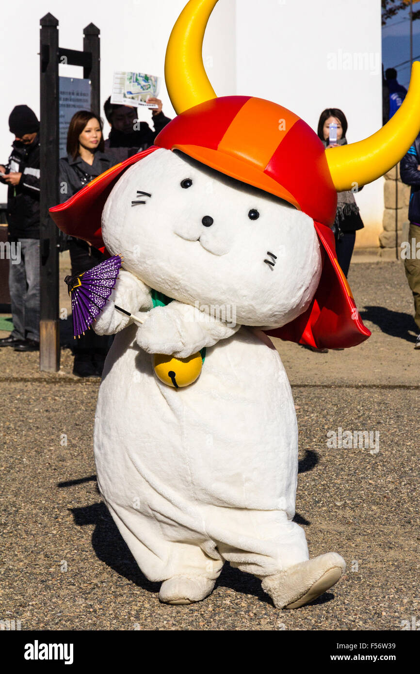 Il castello di Hikone e mascotte della città, gatto bianco, rosso da indossare casco samurai e la campana attorno al collo. Noto come Hikonyan. In piedi rivolta verso il visualizzatore. pubblico dietro. Foto Stock