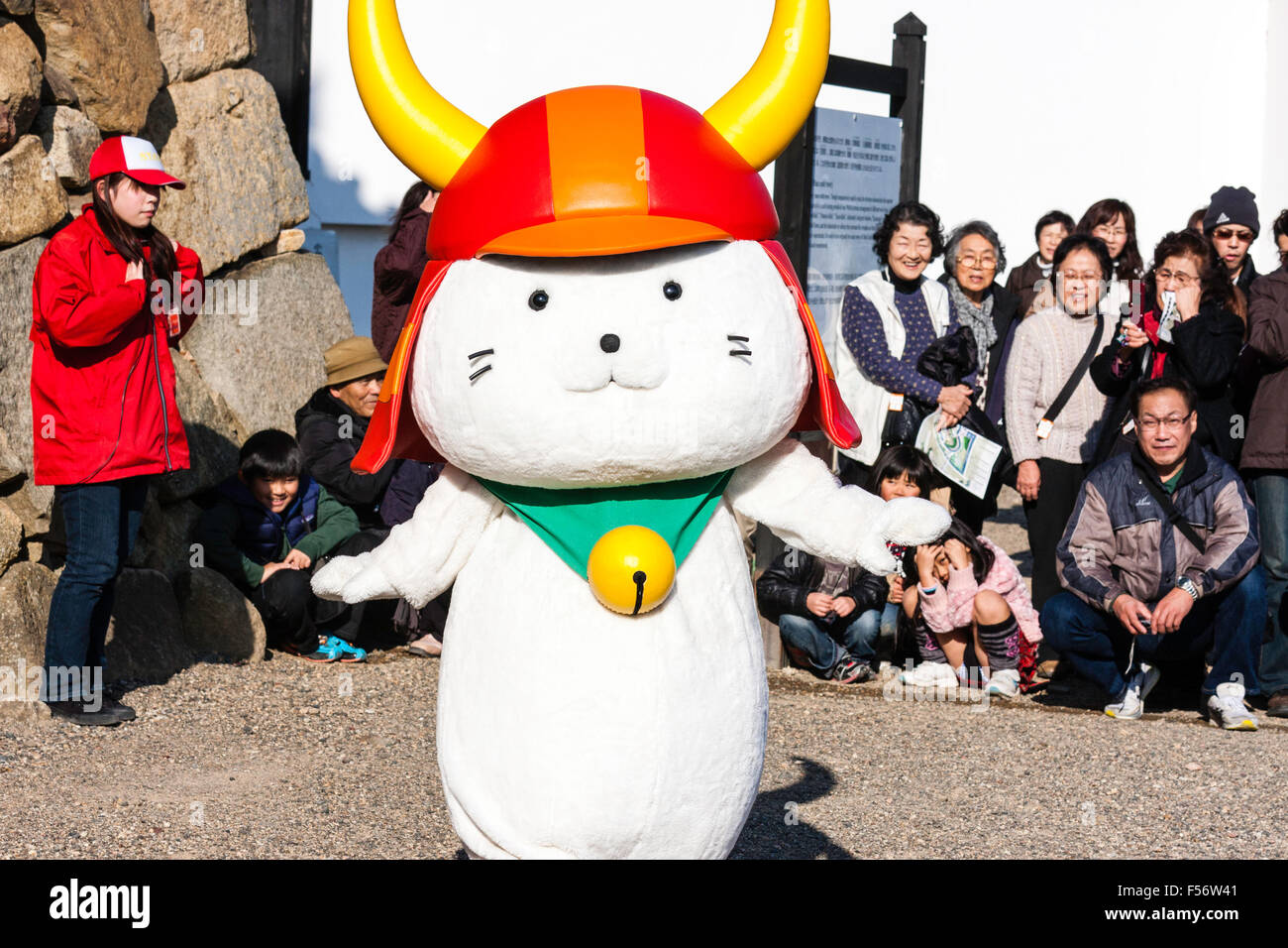 Il castello di Hikone e mascotte della città, gatto bianco, rosso da indossare casco samurai e la campana attorno al collo. Noto come Hikonyan. In piedi rivolta verso il visualizzatore. pubblico dietro. Foto Stock