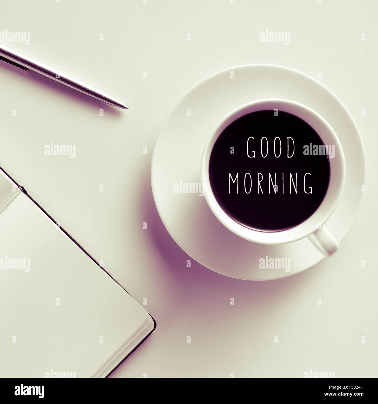 Alta angolazione di un tavolo bianco con una penna, un notebook e una tazza di caffè con il testo di buona mattina scritto in esso Foto Stock
