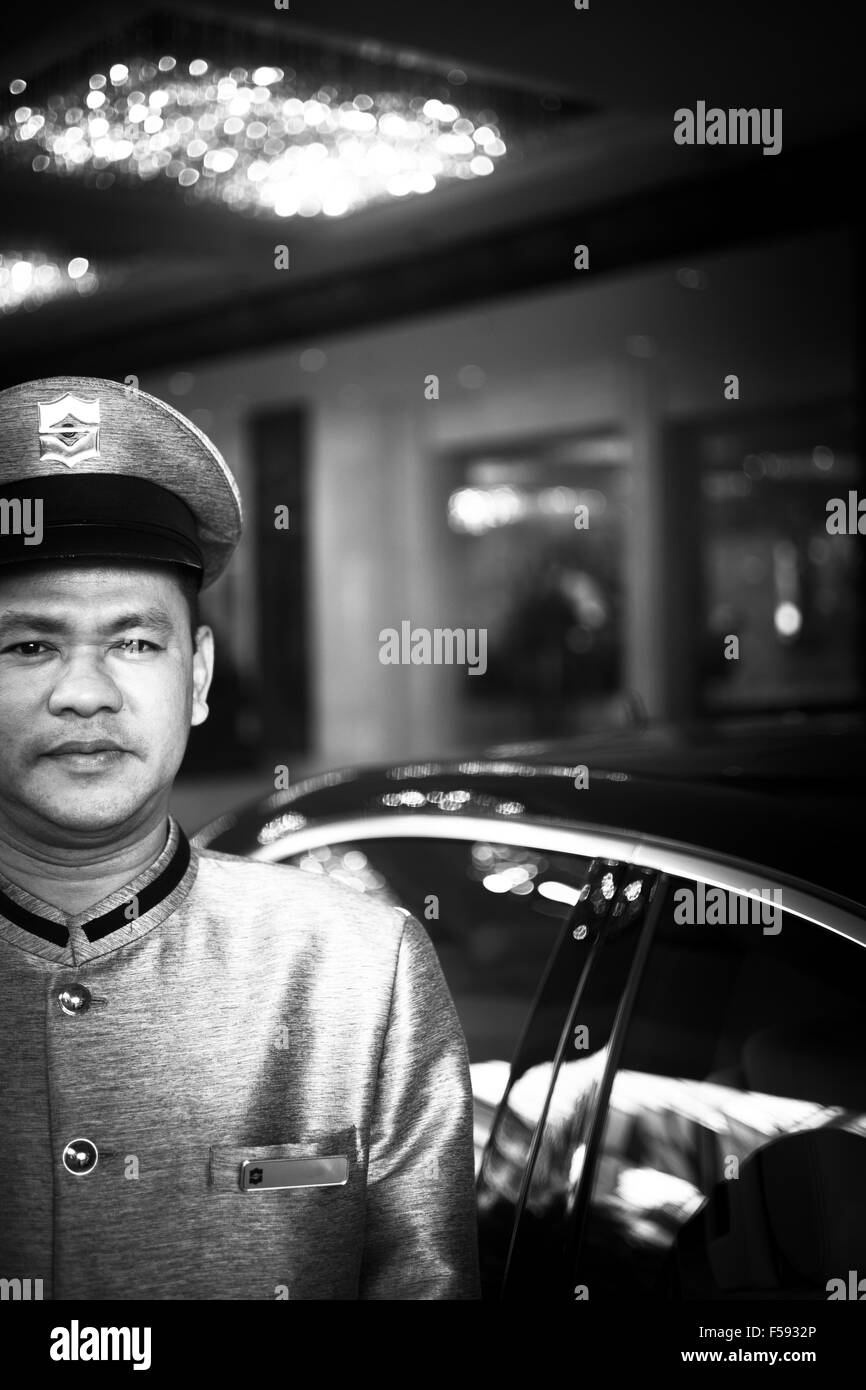Bangkok, Tailandia - 29 Novembre 2013: PARCHEGGIO attendant in piedi al di fuori di lusso a cinque stelle di gran luxe hotel nel centro di Bangkok in Thailandia che indossa la seta Tailandese uniforme. In bianco e nero ad alto contrasto foto digitale di un assistente di parcheggio in posh seta unifo tailandese Foto Stock