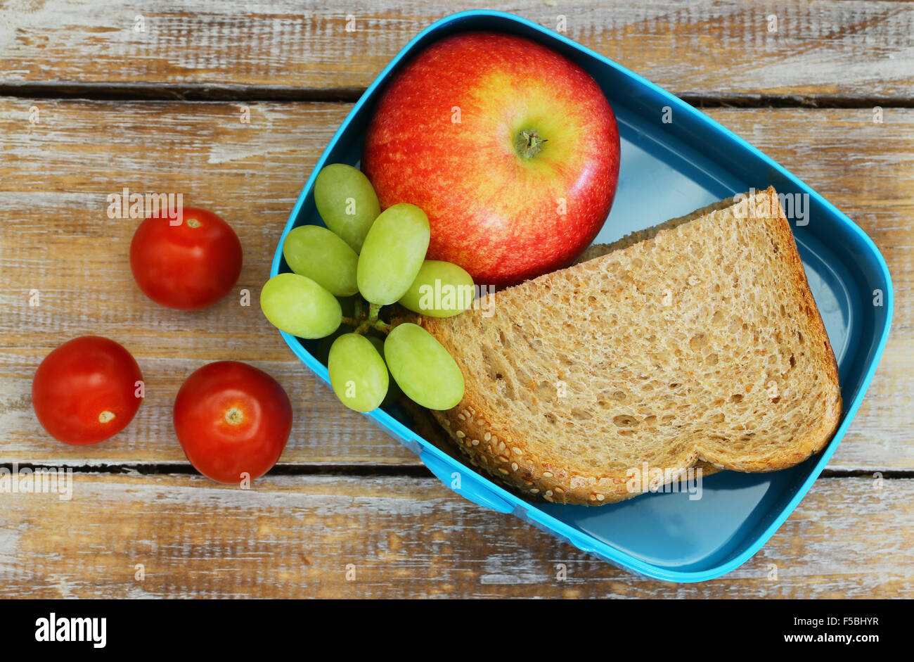 Scatola di pranzo contenenti marrone sandwich di pane, mela rossa, uva e pomodori ciliegini rustico sulla superficie di legno Foto Stock