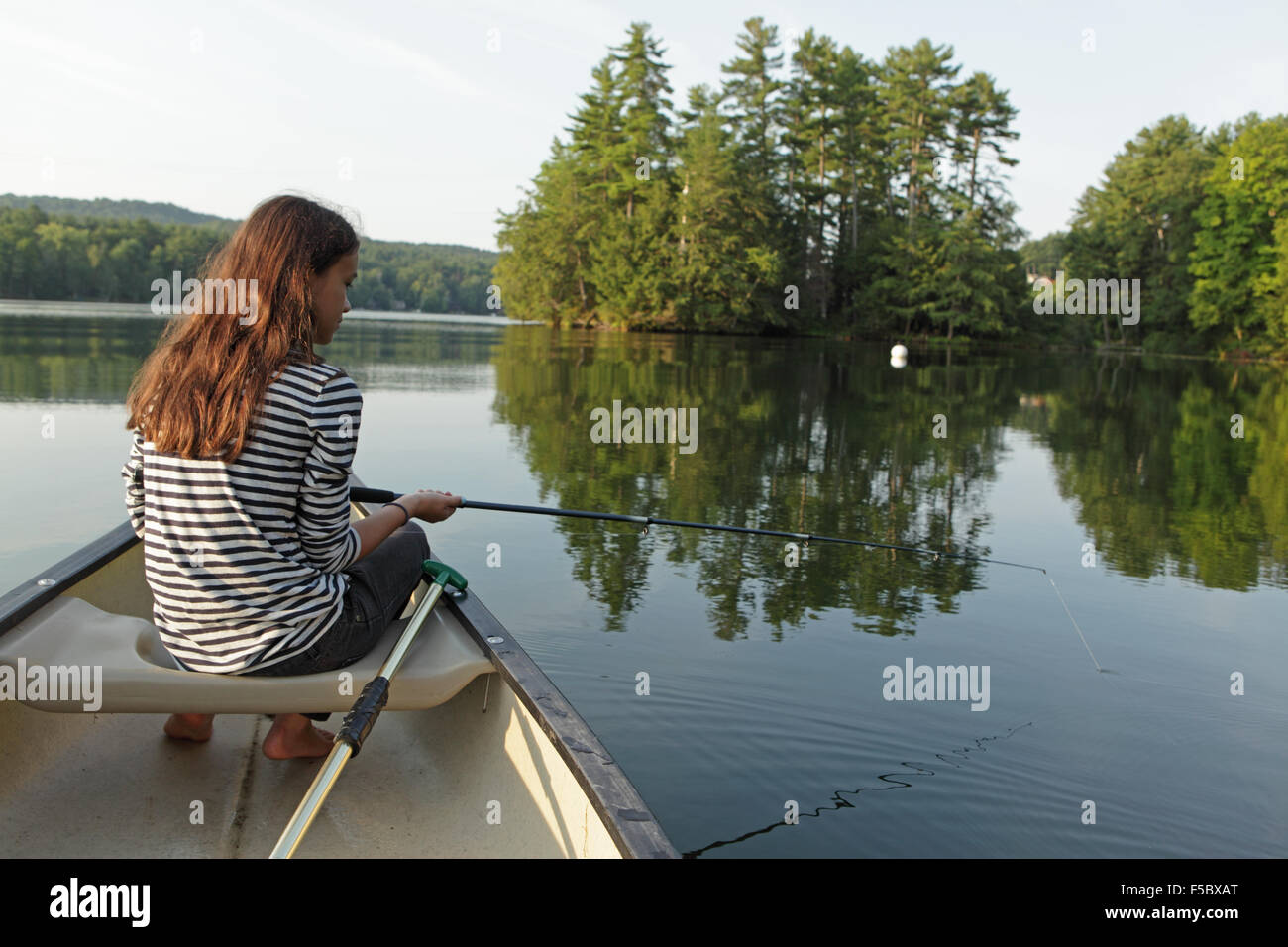 Ragazza giovane la pesca da una canoa su un lago calmo con alberi in background Foto Stock